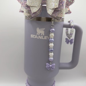 May include: A lavender Stanley tumbler with a decorative bow and bead embellishments. The tumbler has a large handle and a straw. The bow is white and lavender with glitter accents. A beaded charm and a bow adorn the handle.