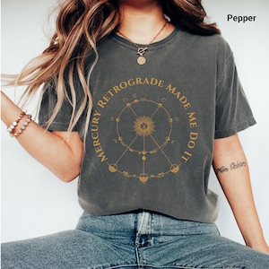 May include: A dark gray t-shirt with the text "MERCURY RETROGRADE MADE ME DO IT" in gold lettering. The design features a circular astrological chart with sun and moon symbols. The shirt is a casual, comfortable style.