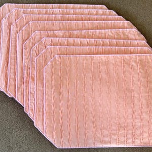 Op de afbeelding: Een stapel van acht perzik-kleurige rechthoekige placemats. Elke placemat heeft een textuur met verticale strepen en een afgeronde achthoekige vorm. De placemats liggen op een neutrale ondergrond.