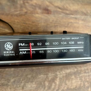 May include: Close-up of a vintage General Electric AM/FM radio dial. The black dial features white lettering indicating FM and AM frequencies, with a red needle pointing to 88 MHz. The radio has a battery backup feature.