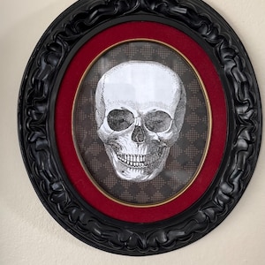 May include: A circular wall art piece featuring a detailed black and white illustration of a human skull centered on a dark checkered background. The artwork is encased in a deep red velvet mat and a thick, ornate black frame with curved edges.