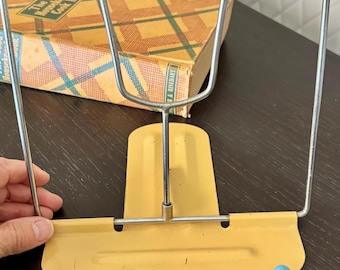 Può includere: Un leggio per ricettari in metallo giallo con una struttura in filo cromato a forma di U per tenere le pagine aperte. La base presenta due piccoli piedini con punte sferiche azzurre. Dietro il supporto si trova un ricettario vintage.