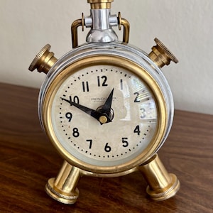 May include: A round Pendu-Lux alarm clock with a metallic silver and brass finish. The clock face is off-white with black Arabic numerals and black hands. It features two brass legs at the base, two side knobs, and a top winding mechanism.