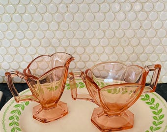 May include: A vintage pink glass creamer and sugar bowl set with handles, on a white and green patterned serving plate. The creamer and sugar bowl have a unique, geometric design. The plate has a green leaf pattern.