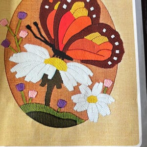 Peut inclure: Une image de référence pour broderie représentant un papillon aux ailes brunes, orange et rouges posé sur des marguerites blanches à cœur jaune. Le motif inclut de petites fleurs violettes et roses sur fond beige, avec le texte "REFERENCE PICTURE" et "8X10".