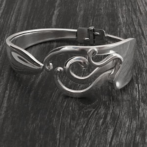 Pu&ograve; includere: Un braccialetto d'argento realizzato con un cucchiaio riutilizzato. Il braccialetto presenta un design decorativo di volute e curve.