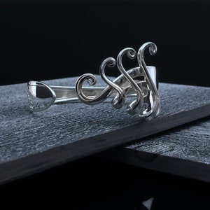 May include: A silver bracelet with a unique, intricate design featuring multiple swirls and curves. The bracelet is resting on a dark grey surface.
