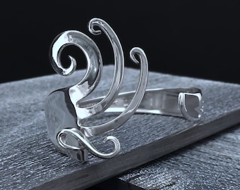 Puede incluir: Pulsera de plata con un diseño abstracto en espiral. La pulsera está hecha de metal y tiene un acabado pulido.