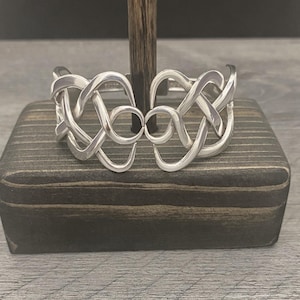May include: A silver Celtic knot bracelet with a woven design. The bracelet is displayed on a dark wooden stand.