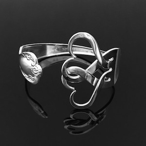 Puede incluir: Un brazalete de plata hecho a partir de una cuchara reutilizada, con forma de dos corazones entrelazados.