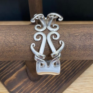 May include: Silver bracelet with a decorative design of swirls and curves. The bracelet is made from a repurposed fork.