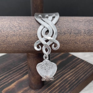 Può includere: Un braccialetto d'argento realizzato con un cucchiaio riutilizzato. Il braccialetto è attorcigliato in un design decorativo e presenta un motivo floreale sul cucchiaio.