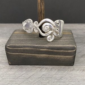 May include: A handcrafted silver bracelet made from repurposed silverware. The bracelet features intricate floral and scrollwork designs, with a spoon bowl and fork tines forming the main structure. The bracelet is displayed on a dark wooden block.