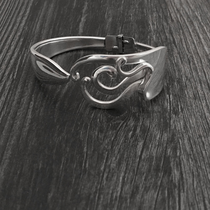 Pu&ograve; includere: Un braccialetto d'argento realizzato con un cucchiaio riutilizzato. Il braccialetto presenta un design decorativo di volute e curve.