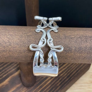 May include: Silver bracelet made from two twisted forks with decorative swirls. The bracelet is displayed on a wooden surface.