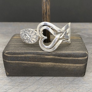 May include: A silver spoon bracelet with a heart design. The bracelet is displayed on a dark wood stand.