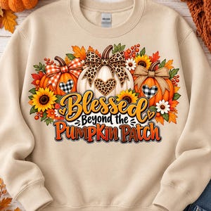 May include: A beige crewneck sweatshirt featuring a graphic print of three pumpkins decorated with bows, sunflowers, and autumn leaves. The text "Blessed Beyond the Pumpkin Patch" is printed in orange and black lettering across the center.