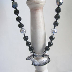May include: A black and silver beaded necklace with a large, gray agate pendant. The pendant has a natural, rough-hewn edge and a large hole in the center.