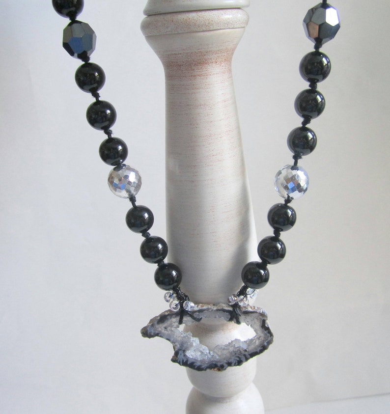 May include: A black and silver beaded necklace with a large, gray agate pendant. The pendant has a natural, rough-hewn edge and a large hole in the center.