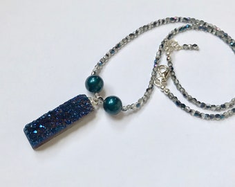 May include: A dark blue crystal pendant necklace with two dark green pearl accents. The necklace is made with a silver chain and small, iridescent beads.