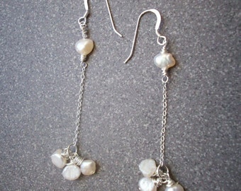 May include: A pair of silver dangle earrings with white pearl accents. The earrings feature a single pearl at the top and a cluster of pearls at the bottom.