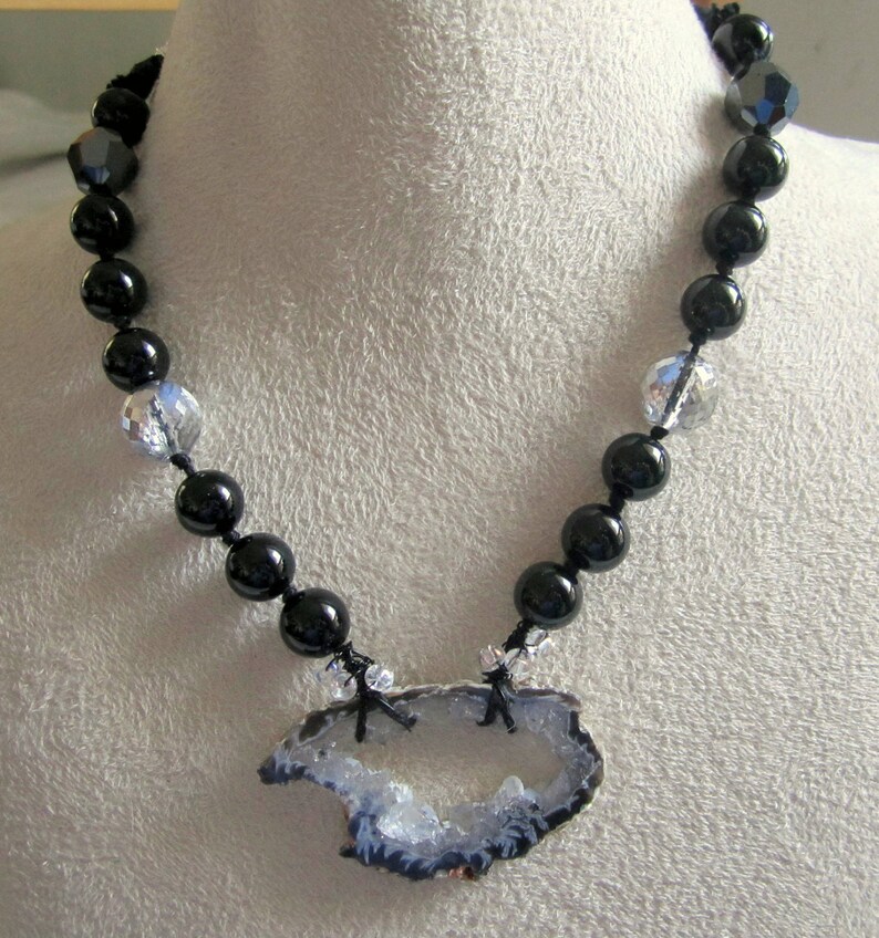 May include: A black beaded necklace with a crystal pendant featuring a large, natural agate slice with a white and gray interior.