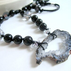 May include: A black and white necklace with a large, grey agate pendant. The pendant has a natural, rough-hewn edge and a large, open center. The necklace is made with black beads and clear glass beads.