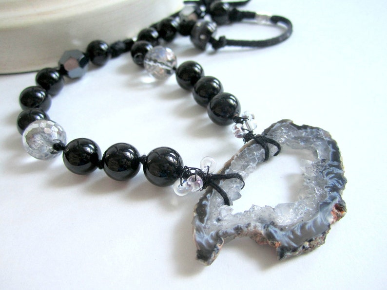 May include: A black and white necklace with a large, grey agate pendant. The pendant has a natural, rough-hewn edge and a large, open center. The necklace is made with black beads and clear glass beads.