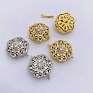 Può includere: Un assortimento di chiusure per gioielli dorate e argentate. Le chiusure sono a forma di fiore e incrostate di cristalli trasparenti. Una chiusura è aperta, rivelando il design interno. È presente anche una piccola chiusura dorata.