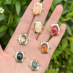 May include: Seven gold-toned rectangular pendants with oval-shaped, colorful gemstones. The gemstones are in shades of peach, cream, red, yellow, orange, teal, and dark blue. Each pendant has small loops on the sides for attaching to jewelry.