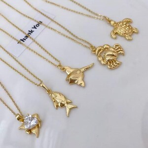 Puede incluir: Collares dorados con varios colgantes, incluyendo una tortuga, un cangrejo, un pez y un colgante en forma de corazón con una piedra transparente. Los collares se muestran sobre una superficie blanca. Las cadenas son delicadas y los colgantes detallados.
