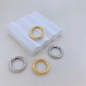 Puede incluir: Cuatro anillos de cierre de resorte metálicos en oro y plata. Los anillos tienen un diseño de cuerda trenzada. Un anillo dorado está sobre una superficie blanca texturizada. Los otros anillos están sobre un fondo blanco.