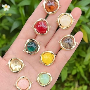 May include: A collection of colorful gemstone cabochons in gold-tone settings. The gemstones are various colors, including red, green, yellow, and pink. Each cabochon has a round shape and is set in a unique, swirling gold frame.