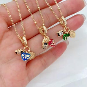 May include: Gold-colored necklaces with charm pendants. Each necklace features a different charm, including a camera, a heart, and a compass. The charms are painted with various colors, including blue, red, green, and gold. The necklaces are displayed on a hand.