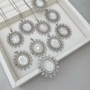 May include: A collection of silver-toned pendant necklaces with oval-shaped, mother-of-pearl centerpieces. Each pendant is surrounded by a decorative, sparkling frame. The necklaces are displayed in a white tray.