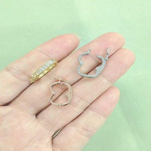 May include: A gold ring with clear stones and two pairs of earrings. One pair is silver and shaped like a heart, and the other pair is rose gold and shaped like an apple. The jewelry is displayed on a hand.