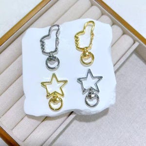 May include: Four metal keychains in gold and silver tones. Two keychains are shaped like a thumbs-up, and two are star-shaped. Each keychain has a ring for attaching keys or other items. The keychains are displayed on a white surface.