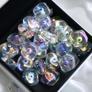 May include: A collection of iridescent glass beads with an evil eye design. The beads are clear with a rainbow sheen and feature a blue eye in the center. They are contained in a black box.