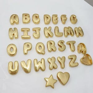 Puede incluir: Una colección de abalorios dorados con letras del alfabeto, junto con un abalorio de estrella y corazón. Las letras están en un estilo de fuente de burbujas, y los abalorios están dispuestos sobre una superficie blanca. Las letras son de la A a la Z.