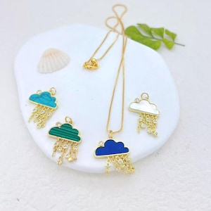 May include: Gold-tone necklaces with cloud pendants in turquoise, green, blue, and white mother-of-pearl. Each cloud pendant has dangling crystal accents, creating a rain effect. The necklaces are displayed on a white, rounded surface.
