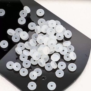 Puede incluir: Un conjunto de pequeñas cuentas de silicona blancas translúcidas, de forma circular y con un agujero central. Las cuentas están dispersas sobre una superficie negra, creando un contraste. Estas cuentas se utilizan probablemente para la fabricación de joyas o artesanía.