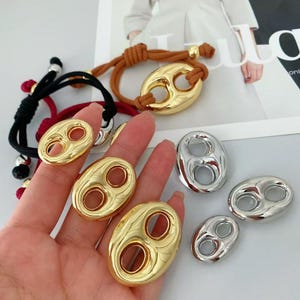 Puede incluir: Varios componentes de joyería dorados y plateados. La imagen muestra piezas ovaladas con dos agujeros circulares, algunas unidas a pulseras. Las joyas se exhiben sobre un fondo claro, con una mano sosteniendo algunas de ellas.