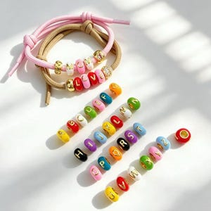 Könnte beinhalten: Zwei elastische Haarbänder in Pink und Beige, jeweils mit goldfarbenen Buchstabenperlen, die MOM und LOVE bilden. Darunter eine Sammlung einzelner runder Perlen in verschiedenen Farben, die jeweils mit einem goldenen Buchstaben von A bis Z versehen sind.