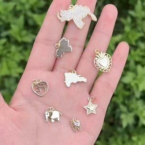 Op de afbeelding: Een verzameling goudkleurige hangers in verschillende vormen, waaronder een ster, een olifant, een hart en een kaart. Sommige hangers zijn versierd met heldere kristallen. De hangers worden getoond tegen een vage groene achtergrond.