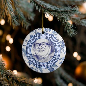 May include: A round ceramic Christmas ornament featuring a portrait of a smiling person with glasses. The ornament has a blue and white floral design with a Greek key border. A gold string is attached for hanging.