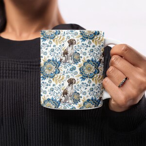 May include: White ceramic mug featuring a repeating pattern of blue and gold floral designs and illustrations of a brown and white dog. The mug has a white handle and is held by a person.