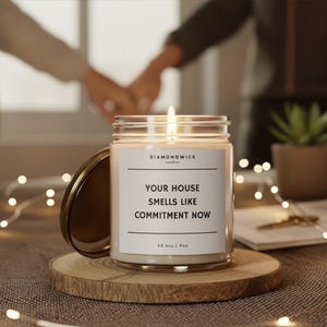 Może przedstawiać: Zapalone świeca w szklanym słoiku ze złotą pokrywką stoi na drewnianej podstawce. Świeca ma białą etykietę z napisem "YOUR HOUSE SMELLS LIKE COMMITMENT NOW". Świeca ma 54g i czas palenia 50 godzin.
