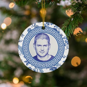 May include: A round white ceramic ornament featuring a blue ink-style portrait of a person in the centre. The portrait is framed by a blue Greek key border, surrounded by a blue and white floral and peacock pattern, hanging by a gold cord.