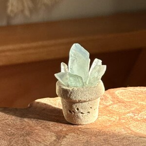 Op de afbeelding: Een kleine witte keramische pot met een tros lichtblauwe glazen kristallen erin. De pot staat op een houten oppervlak.