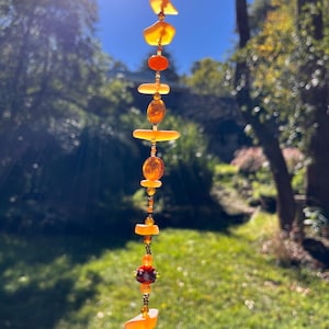 May include: A decorative hanging strand featuring amber-toned beads and stones. The beads are of various shapes and sizes, strung on a thin, gold-coloured wire. The background is a blurred outdoor scene with trees and greenery, suggesting a sunny day.